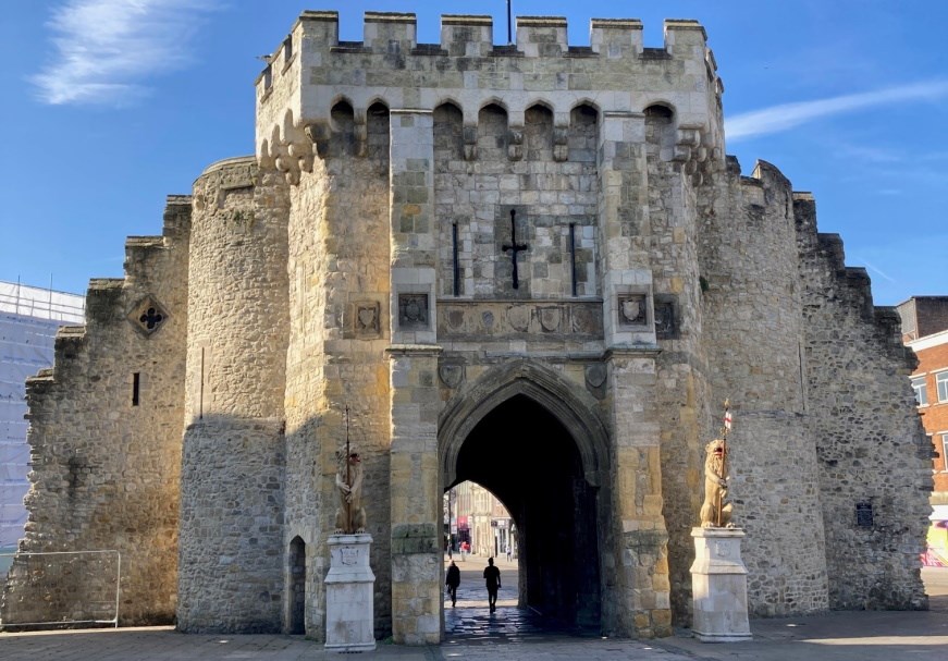 Image of medieval stone and flint monument with arrow slit windows and an archway in the middle. Two gold lions statues are in front of the monument holding two flags.