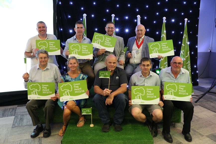 Image of people sat down smiling and holding Green Flag Award 2023/24 plaques and flags. Sat in front of a black screen with white lights and five Green Flag Award flags. Artificial grass is laid on the floor.