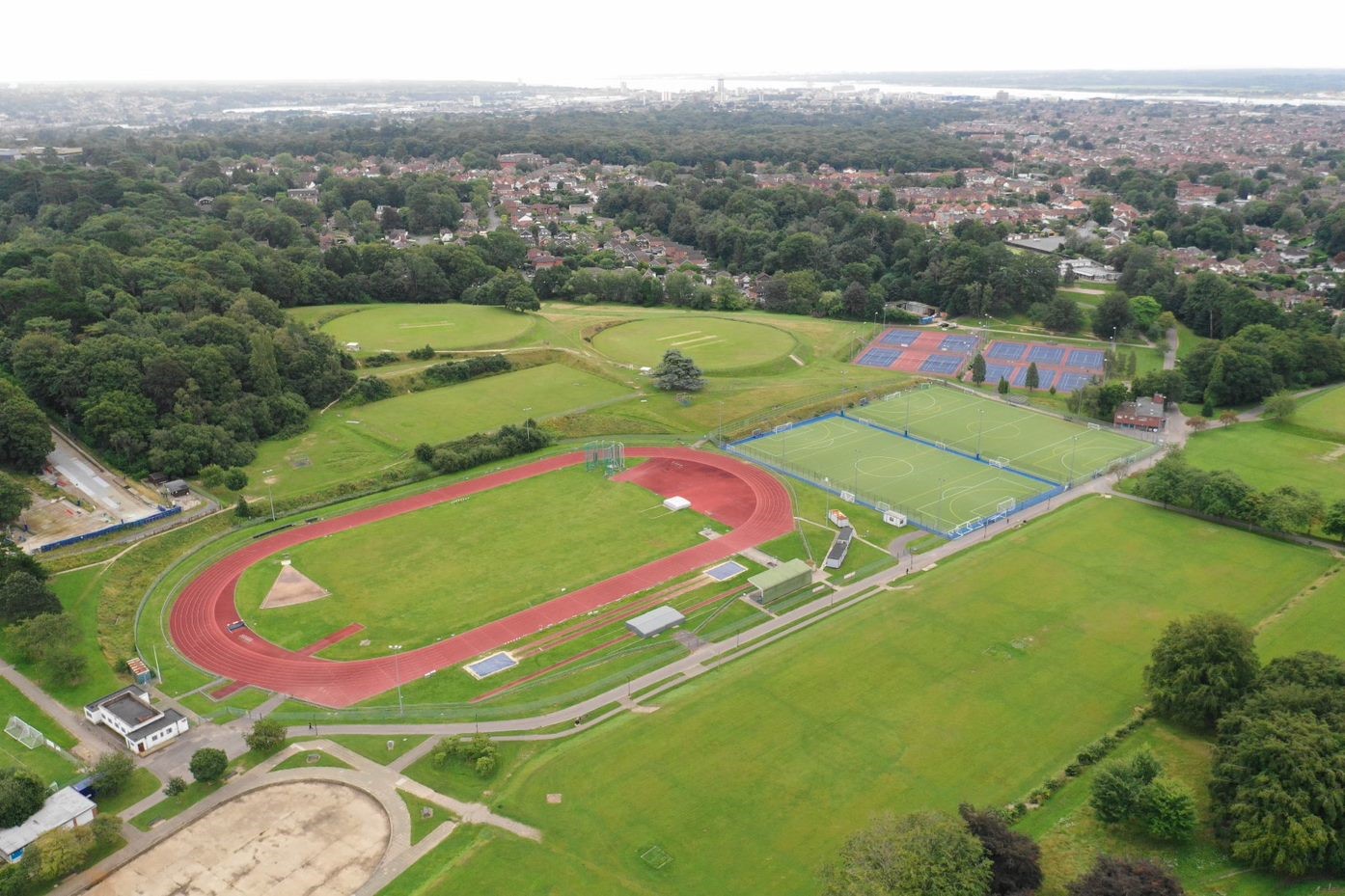Drone aerial image of sports centre with burgundy athletics track, a white building, two green football pitches and a burgundy and blue tennis court in the distance. The facilities are surrounded by lots of green space and trees.