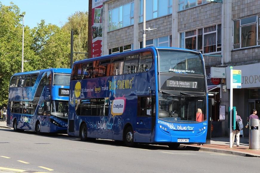 A photo of two Bluestar buses at the bus stops on Above Bar Street, with the shops behind them and the edge of the park in the background.