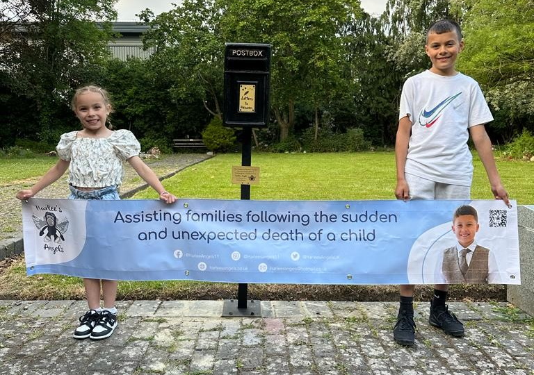 Louie and Elsie-Mae Steele with post box. They hold a banner for Harlee's Angels which says "Assisting families following the sudden and unexpected death of a child".