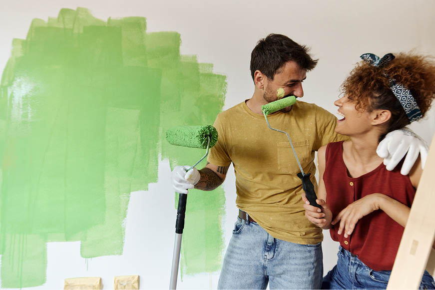 A man and woman painting a wall. The woman has daubed some paint on the man's face and they are both laughing