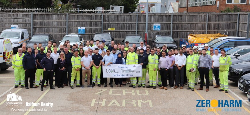 A group photo of Southampton City Council and Balfour Beatty staff in a car park. Roughly 80 people. They are wearing either office or high-visibility clothing. They hold a banner saying "Recognising 1,000 days. 1 million hours. Lost Time Injury (LTI) free". Line painting on the ground says "Zero Harm". Logos on the image: Southampton City Council, Balfour Beatty, Working in partnership, Zero Harm - Make Safety Personal.