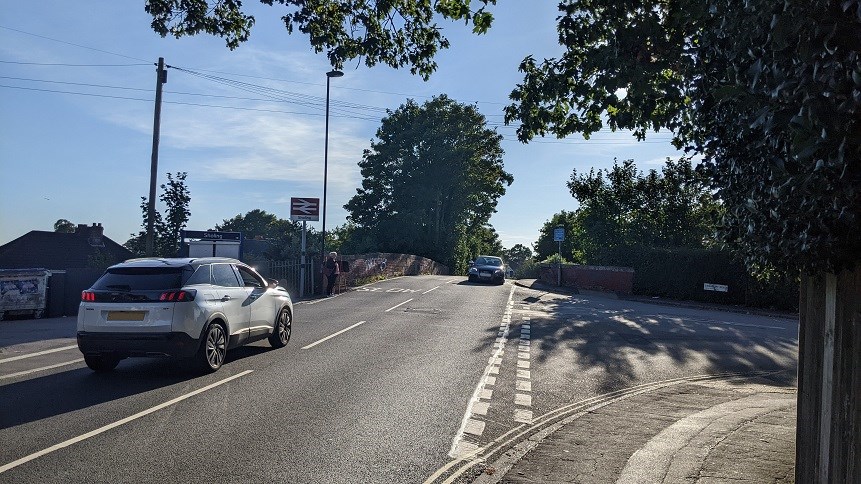 A photo of the junction of Station Road and Cranbury Road in Sholing. A white 4x4 car drives away from the photographer, while a dark car comes forward over the railway bridge. Trees are in the background and to the right of the photo, while to the left, beyond the white car, is the sign for Sholing Station and a pedestrian waits at the Station entrance to cross the road.