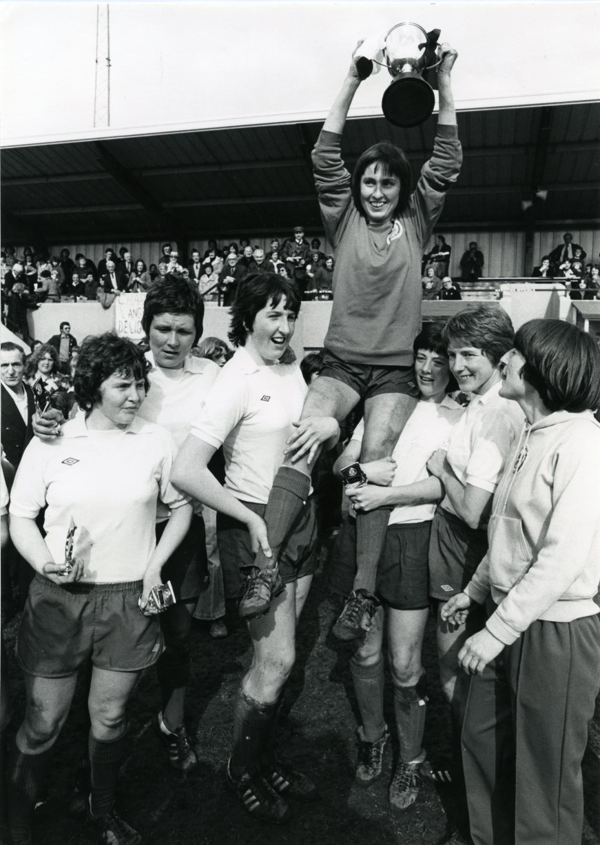 Women's football team celebrating with their trophy. One of the players is held aloft.