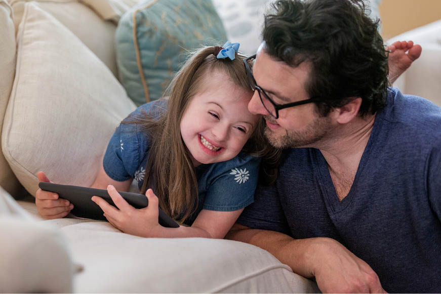 A smiling child using a tablet computer on the sofa with her father
