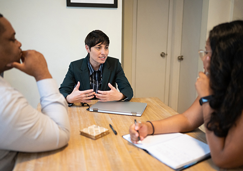 Three people sitting at a table in a home. One woman is explaining something to the other two people. 