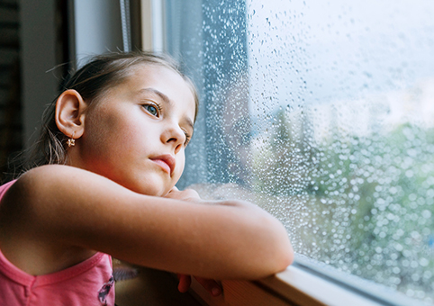 A sad looking girl looking out of a window with water droplets on it