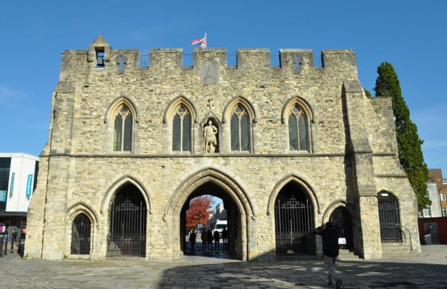 Image of stone medieval building with arrow slit windows and, four archways with black gates and another archway with people walking through with blue skies in the background