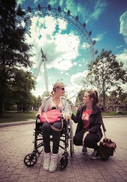 A person in a wheelchair talking to someone kneeling beside them. There is a Ferris wheel behind them and it is a sunny day