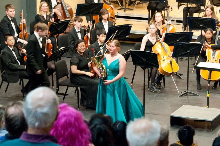 The Youth Orchestra largely seated in front of an audience. A soloist is standing in front of the orchestra
