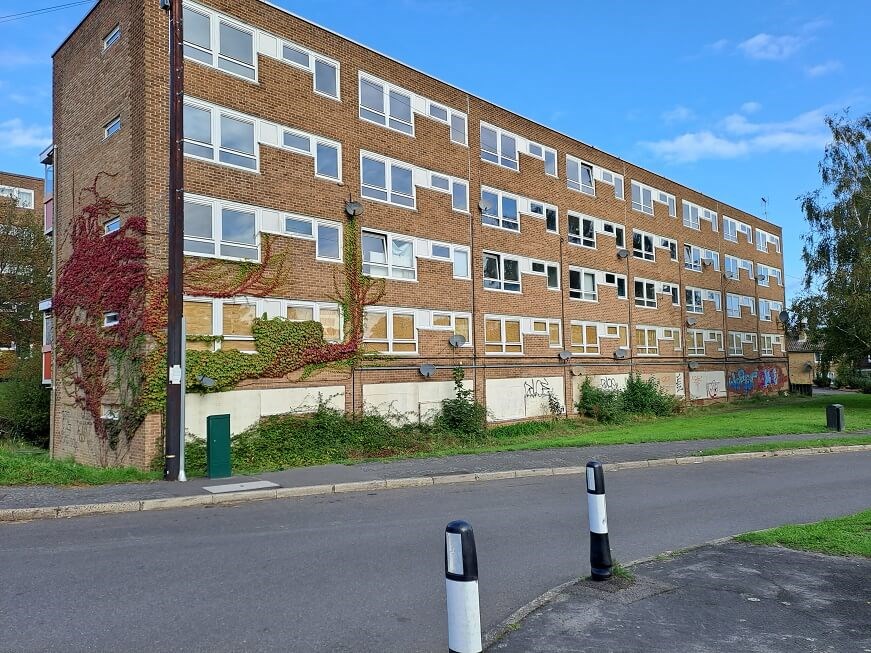 Image of brown block of flats with white windows and white garages at the bottom. Green bushes, grass surround the block. At the side of the block is red and green moss growing.