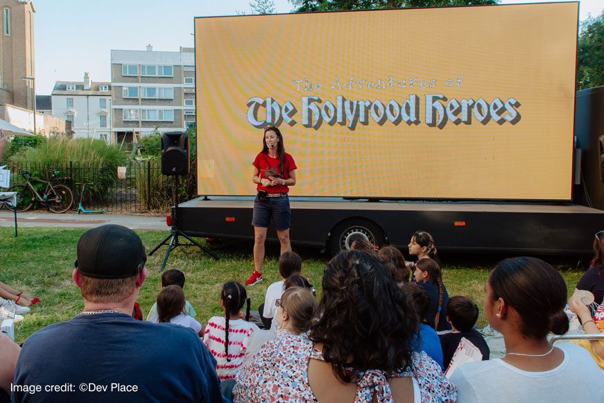 A Crowd Looking At A Woman Standing In Front Of A Screen That Says: The Adventures of The Holyrood Heroes. Image credit: copyright Dev Place A Crowd Looking At A Woman Standing In Front Of A Screen That Says: The Adventures of The Holyrood Heroes. Image credit: copyright Dev Place