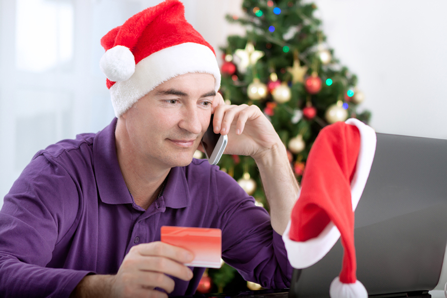 Man Wearing Xmas Hat Using A Mobile Phone Whilst Looking At Laptop And Holding Credit Card 871X581
