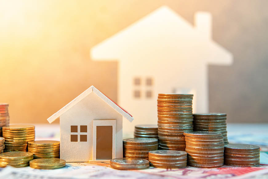 A small wooden house sitting on bank notes, surrounded by stacks of coins. A larger house of the same style is in the background