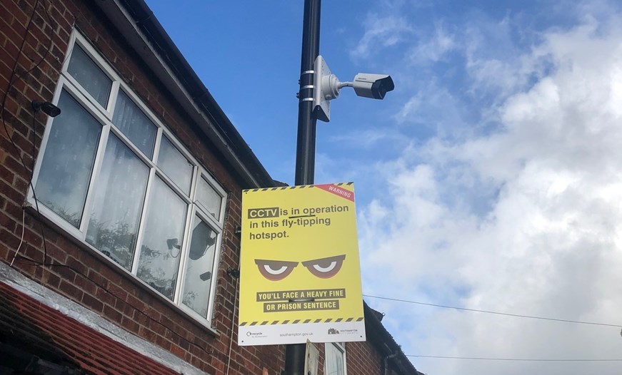 Yellow and black warning sign underneath a CCTV camera. Text: 'CCTV is in operation in this fly-tipping hotspot. You'll face a heavy fine or a prison sentence'